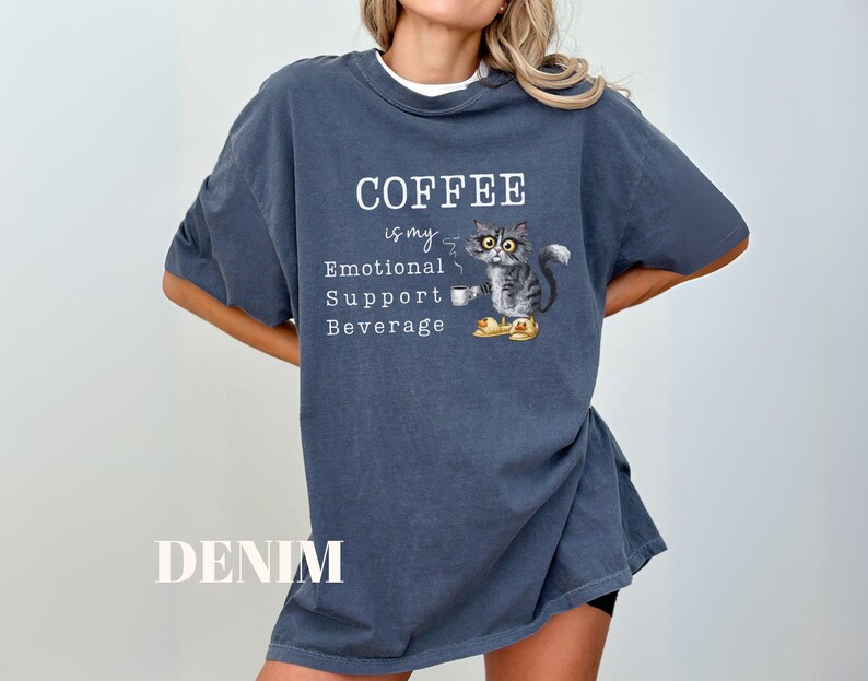May include: Denim blue t-shirt with the text "COFFEE is my Emotional Support Beverage." The shirt features a graphic of a cat holding a coffee cup. The cat is wearing yellow slippers. The word "DENIM" is printed at the bottom.