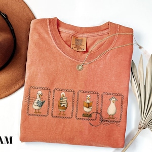 May include: Coral-colored Comfort Colors t-shirt featuring a whimsical design of four cartoon geese in postage stamp frames, each engaged in reading. A gold necklace and a brown hat are also visible. The shirt has a crew neck and short sleeves.