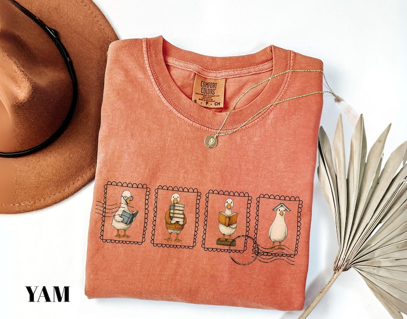May include: Coral-colored Comfort Colors t-shirt featuring a whimsical design of four cartoon geese in postage stamp frames, each engaged in reading. A gold necklace and a brown hat are also visible. The shirt has a crew neck and short sleeves.