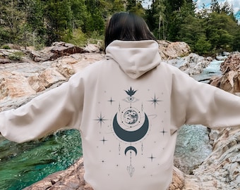 May include: Beige hoodie with a celestial design on the back. The design features a crescent moon, stars, an eye, and other celestial elements in a dark teal color. The hoodie has a hood and long sleeves.