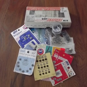 May include: A collection of sewing notions including a Dot Snappers kit box, various cards of metal snap fasteners from brands like Scovill and Prims, and small clear plastic containers holding loose silver-toned metal snaps and fasteners.