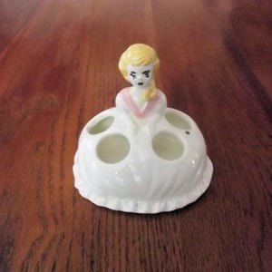 May include: A white ceramic toothbrush holder shaped like a woman in a dress. The woman has blonde hair and a pink accent on her dress. The holder has four circular openings for toothbrushes.