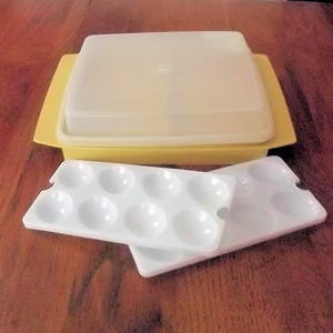 May include: A yellow rectangular container with a lid, and two white, oval-shaped paint palettes. The container has a yellow base and a translucent lid. The palettes have multiple circular wells for holding paint.