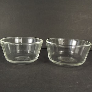 May include: Two clear glass bowls with a slightly angled design. The bowls are transparent, allowing visibility of their contents. They are set against a dark background, highlighting their shape and clarity. Ideal for serving small portions.