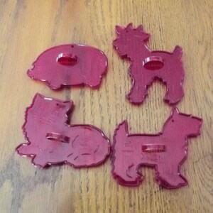 May include: A set of four translucent dark red plastic cookie cutters. The shapes include a pig, a standing dog, a sitting dog, and another standing dog. Each cutter features a small circular handle on the top surface for easy gripping.