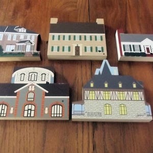 May include: Five wooden house-shaped decorative items. Each house features a unique architectural design, including brick, siding, and various window styles. The houses are painted in a variety of colors, including beige, gray, and red, and are arranged on a wooden surface.