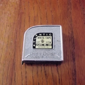 May include: A silver-colored Atlantic White Tape measuring tape. The tape measure has black text that reads "WHITE TAPE", "12FT", and "ADD 2 INCHES". The tape measure is on a wooden surface.