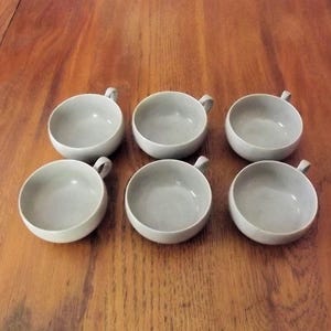 May include: A set of six matching light gray ceramic bowls, each featuring a smooth, rounded basin and a small, singular handle extending from the side. The bowls are arranged in two rows of three, showcasing a uniform, minimalist design.