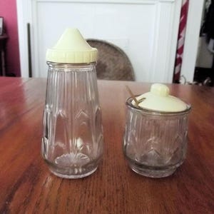 May include: Two vintage glass containers with cream-colored lids. One is a tall, fluted shaker, and the other is a small jar with a spoon. Both have a decorative pattern and are likely for kitchen use.
