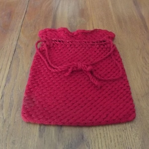May include: A handmade red crocheted drawstring bag. The bag has a textured pattern and a decorative bow closure. The top edge is scalloped. The bag is sitting on a wooden surface.