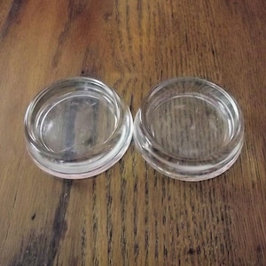 May include: Two clear glass coasters with a circular design. The coasters have a slightly raised edge and a flat center. They are sitting on a wooden surface with a natural wood grain pattern.