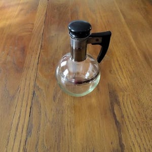 May include: A clear glass decanter with a round body, a silver-colored metal neck, and a black handle and stopper. The decanter sits on a wooden surface.