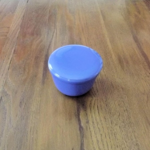 May include: A small, round, lavender-colored container with a matching lid. The container is sitting on a wooden surface. The container is likely used for storage or display.