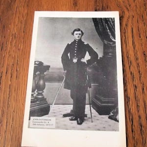 May include: Black and white photograph of a man in a military uniform, standing with one hand on his hip and holding a sword. The photograph includes the text "JOHN PATTERSON Commander Co. A 20th Infantry, 1872-77."