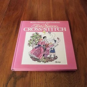 May include: A pink book titled "Better Homes and Gardens Four Seasons Cross-Stitch" with a colorful illustration of a family flying kites in a spring setting. The book features the word "Spring" at the bottom of the cover.