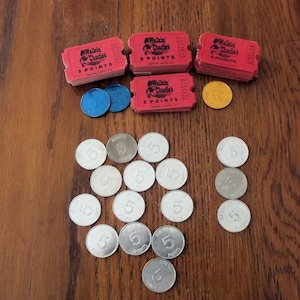 May include: An assortment of vintage game tokens and tickets on a wooden surface. The image includes stacks of red "Walkin' Charlie's 5 Points" tickets, blue and gold tokens, and silver tokens with the number "5" printed on them.