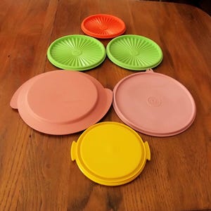 May include: A collection of colorful plastic Tupperware lids in various sizes and shapes. The lids are in shades of orange, green, pink, and yellow. Some lids have a ridged design, while others are smooth. The lids are arranged on a wooden surface.