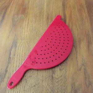 May include: A bright red, semi-circular colander featuring a flat, perforated surface with numerous small, evenly spaced drainage holes. The kitchen tool includes an integrated handle with a circular cutout at the end for convenient hanging storage.