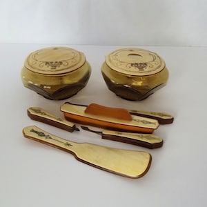May include: A vanity set featuring two amber-colored glass jars with round lids, a long-handled brush, a smaller brush, and a metal tool. Each piece displays a cream-colored inlay with a decorative black line drawing of a floral basket motif.