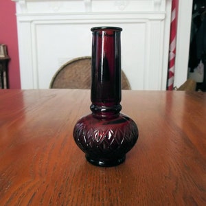 May include: A ruby red glass vase with a bulbous base and a tall, cylindrical neck. The base features a diamond pattern, and the vase sits on a wooden table. The vase is approximately 10 inches tall.