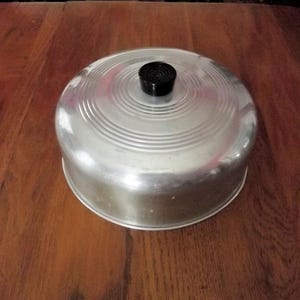 May include: A round, silver-colored metal food cover with a smooth, polished finish and concentric circular ridges on the top surface. The cover features a small, black, cylindrical knob handle centered on the top for easy lifting and placement.