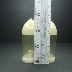 May include: A clear, plastic container with a cream-colored top and base, approximately 3 inches tall. A ruler is placed next to the container for measurement. The container has a small, cream-colored spout at the bottom.