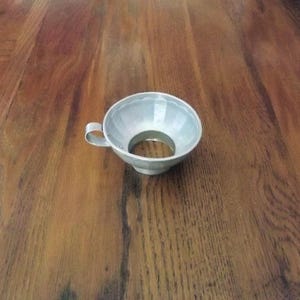 May include: A silver-colored metal canning funnel with a handle. The funnel has a wide, fluted rim and a large opening at the bottom. It is designed to fit over the mouth of a jar for easy filling. The funnel is sitting on a wooden surface.