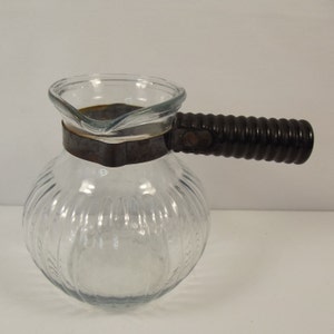 May include: Clear glass coffee pot with a ribbed body and a dark brown, ridged handle. The pot has a metal band around the middle and a spout for pouring. The handle is attached with a metal bracket.