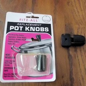 May include: A black replacement pot knob by Fitz-All in its original white and pink retail packaging. The packaging features text including Replacement Pot Knobs, Heat Resistant, and Dishwasher Safe. A second identical black knob sits beside it.