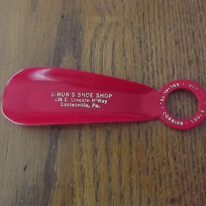 May include: A red shoe horn with a loop handle. The shoe horn has the text "SIMON'S SHOE SHOP 238 E. Lincoln H'Way Coatesville, Pa." printed on it. The handle has the text "RED SOLES SOCIALITES COBBIES" printed on it.