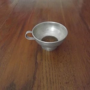 May include: A small, silver-colored metal funnel with a handle. The funnel has a wide, circular opening and a narrower base. The handle is a small, curved piece of metal attached to the side of the funnel. The funnel is sitting on a wooden surface.