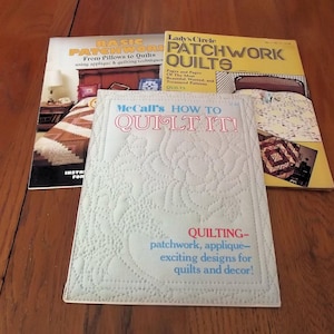 May include: Three vintage quilting books are displayed on a wooden surface. The books include "Basic Patchwork," "Lady's Circle Patchwork Quilts," and "McCall's How to Quilt It!" The covers feature images of quilts and text about quilting techniques.
