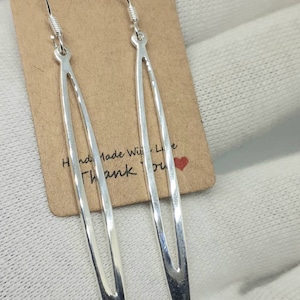 May include: A pair of silver dangle earrings with a long, narrow, oval shape. The earrings have a hammered texture and a pointed tip. The earrings are hanging on a brown card with the text "Handmade With Love Thank You" and a red heart.