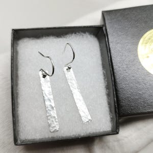 Può includere: Un paio di orecchini a barra argentati con una texture martellata, presentati in una confezione regalo nera con imbottitura bianca. Gli orecchini hanno un design semplice e moderno con chiusura a gancio.