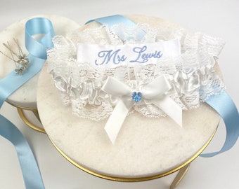 May include: White lace garter with a white satin bow and a blue rhinestone. The garter has a white satin ribbon with the text "Mrs. Lewis" embroidered in blue.