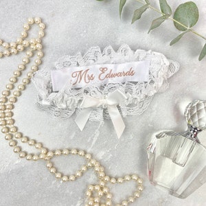May include: White lace garter with a satin ribbon that says "Mrs. Edwards" in gold embroidery. The garter is on a white surface with a pearl necklace and a clear glass bottle of perfume.
