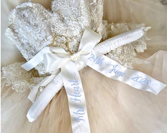 May include: A white padded clothes hanger featuring a gold metal hook and a white satin ribbon tied in a bow. The ribbon is embroidered with the blue text "Mrs Heart" and "24th June 2028". The hanger is positioned over a white lace garment.