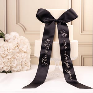 May include: A three-tiered white cake on a clear glass pedestal stand, adorned with a large black satin ribbon tied in a bow at the top. The ribbon features white cursive text reading "Theo & Lily" on one side and "It was always you" on the other.