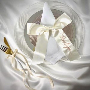 Puede incluir: Una servilleta de tela blanca doblada sobre un plato de sitio de vidrio transparente, atada con una cinta de sat&eacute;n color crema con el nombre Penelope en letra cursiva. Un juego de tenedor y cuchillo dorados est&aacute; atado con una cinta a juego.