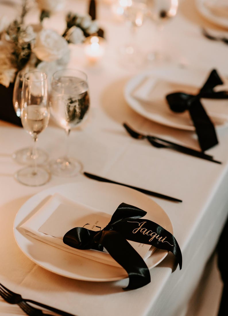 Puede incluir: Un plato blanco con una servilleta blanca doblada y un lazo de sat&eacute;n negro. La cinta presenta el nombre Jacqui escrito en una elegante caligraf&iacute;a blanca. Un tenedor y un cuchillo negros est&aacute;n colocados junto al plato sobre un mantel blanco.