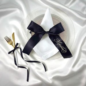 May include: A white cloth napkin folded into a triangle on a clear glass charger plate, tied with a black satin ribbon featuring the name Marina in white script. A gold fork and knife are tied together with a matching black satin ribbon.