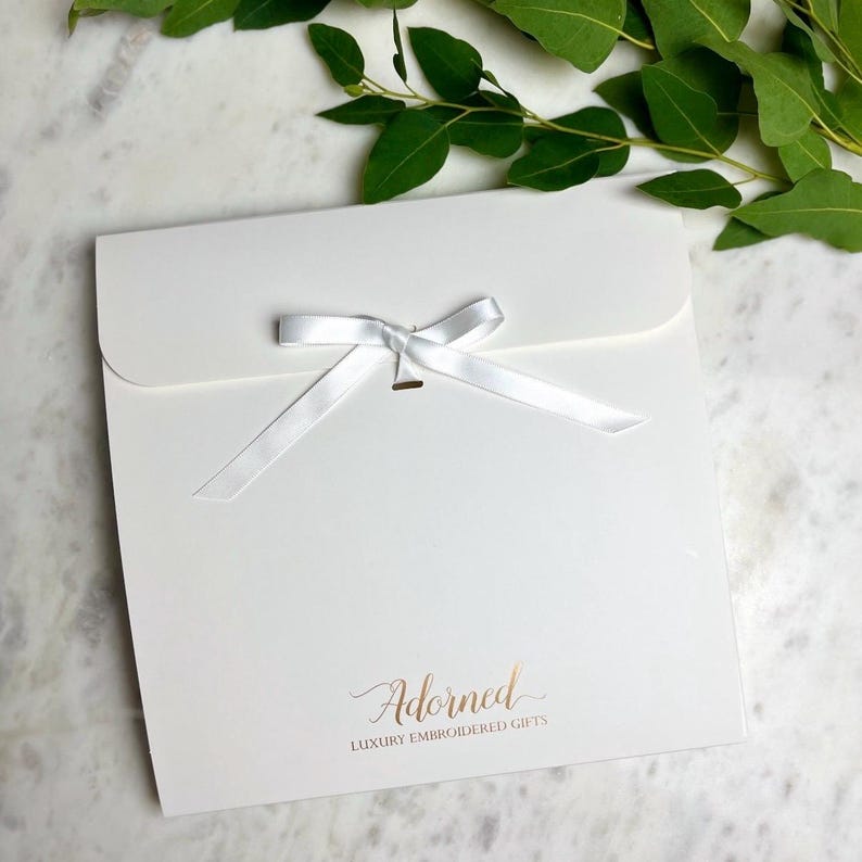 May include: A square white gift envelope featuring a centered white satin ribbon tied in a bow. The bottom center displays the text Adorned LUXURY EMBROIDERED GIFTS in a metallic copper font. The envelope has a curved flap closure at the top.