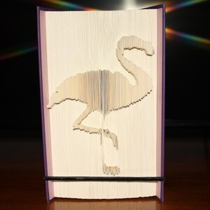 Peut inclure: Un livre dont les pages sont pliées pour créer une sculpture 3D d'un flamant rose. Le livre est debout sur une surface en bois.