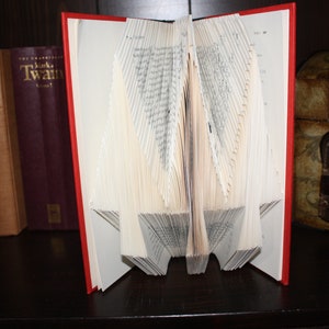 Peut inclure: Un livre relié rouge avec les pages pliées en forme de 3D ressemblant à un oiseau. Le livre est sur une étagère brun foncé avec d'autres livres et une boîte en bois.