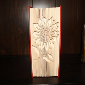 Puede incluir: Un libro rojo con un diseño de girasol recortado en papel blanco. El libro está de pie sobre un estante de madera.