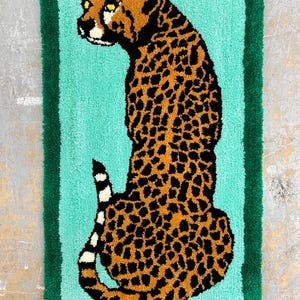 May include: A rug with a green border and a turquoise background features a brown and black cheetah with yellow eyes. The cheetah is facing left and is sitting on its haunches.