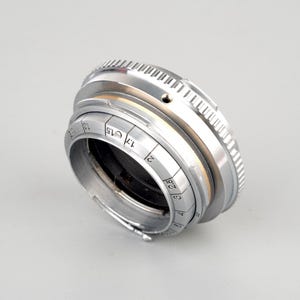 Op de afbeelding: Een zilverkleurige cameralens met een geribbelde focusring en diafragma-instellingen. De lens heeft een reeks getallen en markeringen die brandpuntsafstanden aangeven. De lens is schuin geplaatst en toont de metalen constructie en het ontwerp.