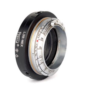 Puede incluir: Un adaptador de lente de cámara negro y plateado con el texto "L39-NEX Rusnild®" impreso en el lateral. El adaptador tiene un anillo plateado con marcas para los ajustes de apertura.