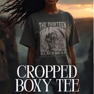 以下が含まれることがあります： ダークグレーのクロップドボックスTシャツ。前面には「THE THIRTEEN」と「BLACKBEAK」の文字が入ったグラフィック。モデルはブルージーンズを着用。背景は夕焼けです。
