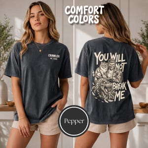 May include: A charcoal gray short-sleeve t-shirt with a small white logo on the front chest reading CRAWLER No. 4122. The back features a large graphic of a person, a cat, and a dinosaur with the text YOU WILL NOT BREAK ME in white lettering.